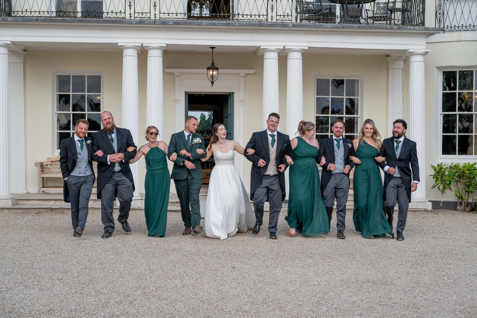 Bridal party arm-in-arm laughing with Rockbeare Manor as backdrop