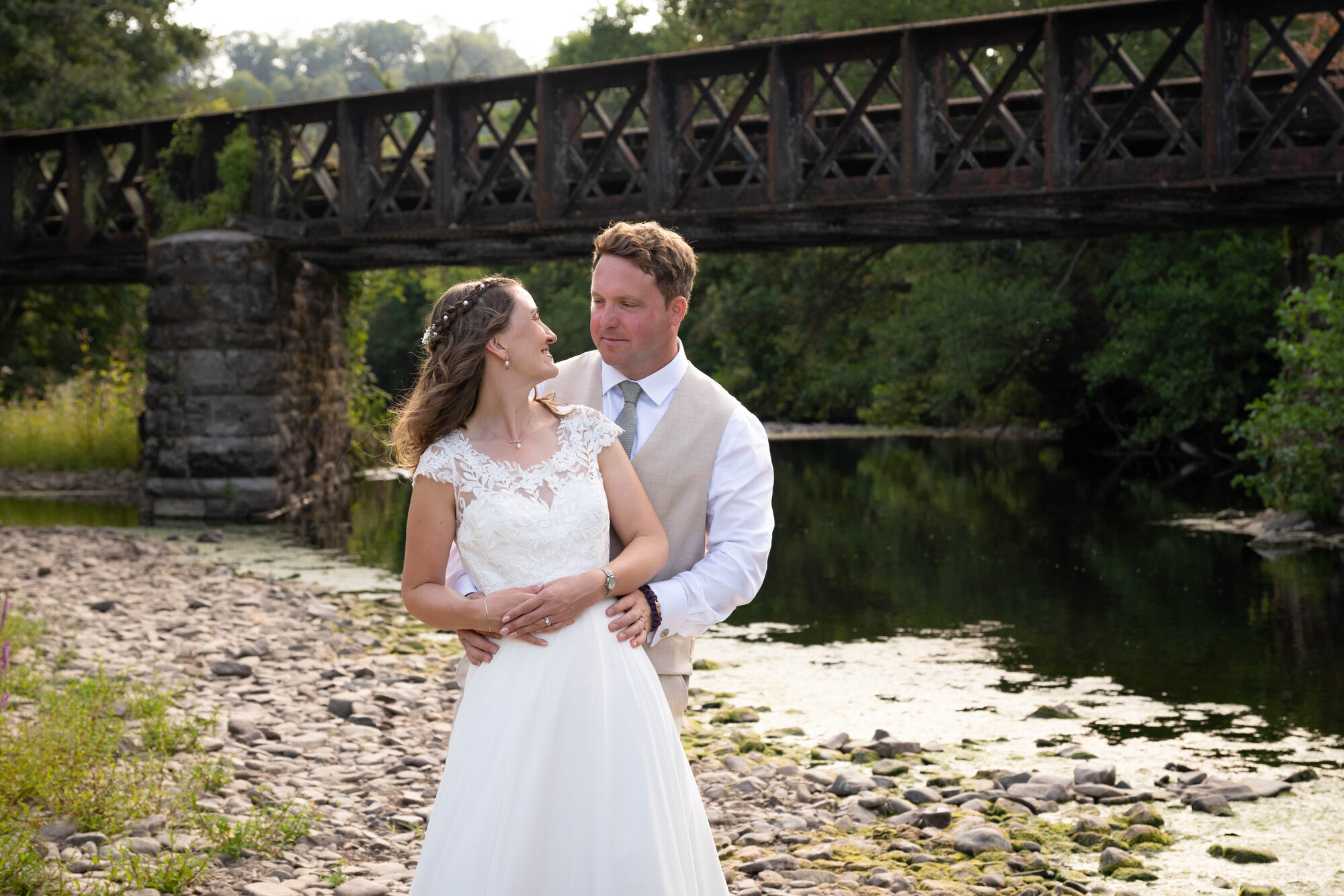 Bride and groom on the riverbank at sunset with Bickleigh Bridge behind them