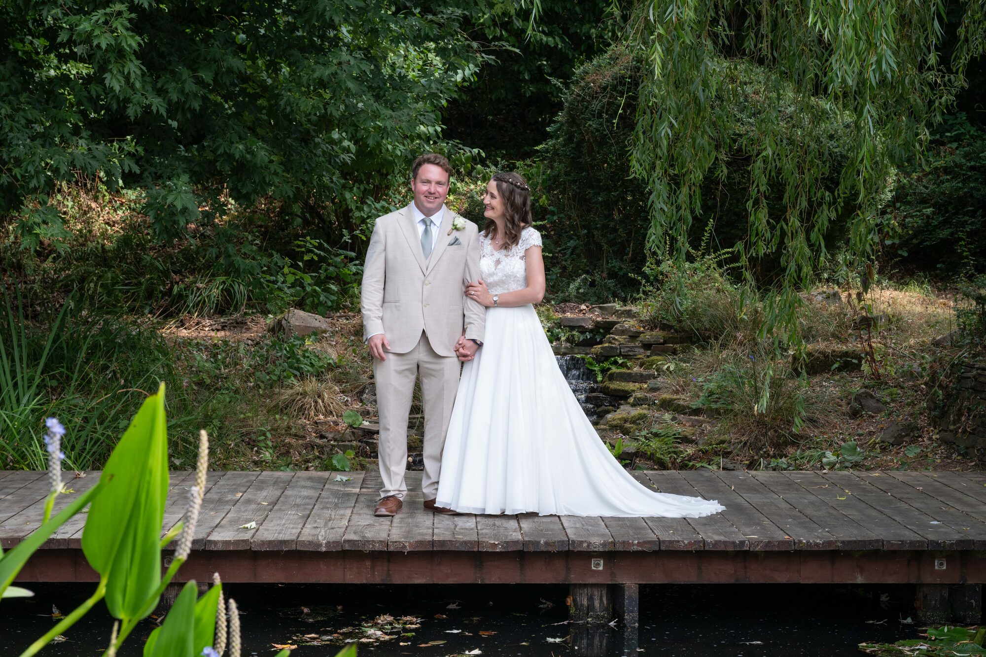 Bride and groom standing on the wooden jetty by the pond with willow tree in the background